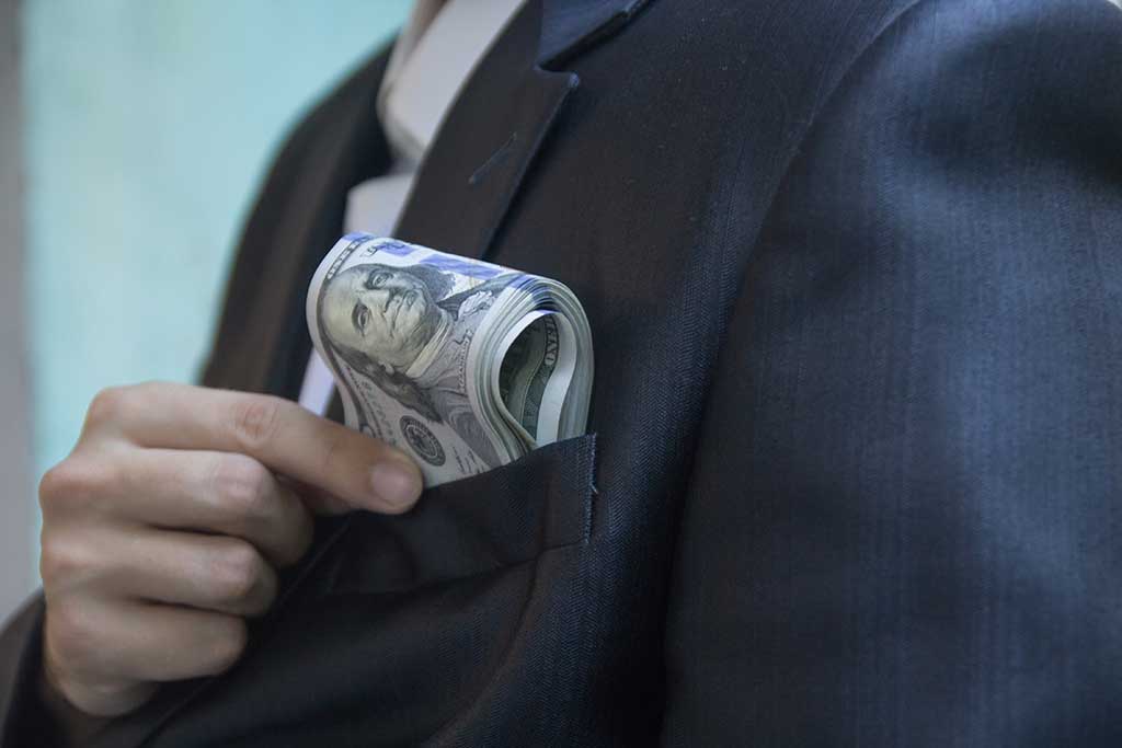 A man in a jacket or a tuxedo puts a bundle of hundred-dollar bills in his breast pocket, close up